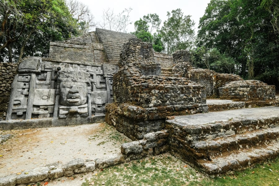 The Mask Temple of Lamanai, an ancient Mayan temple featuring prominent stone masks with detailed carvings of faces, representing deities or rulers, set within a lush jungle environment in Belize.